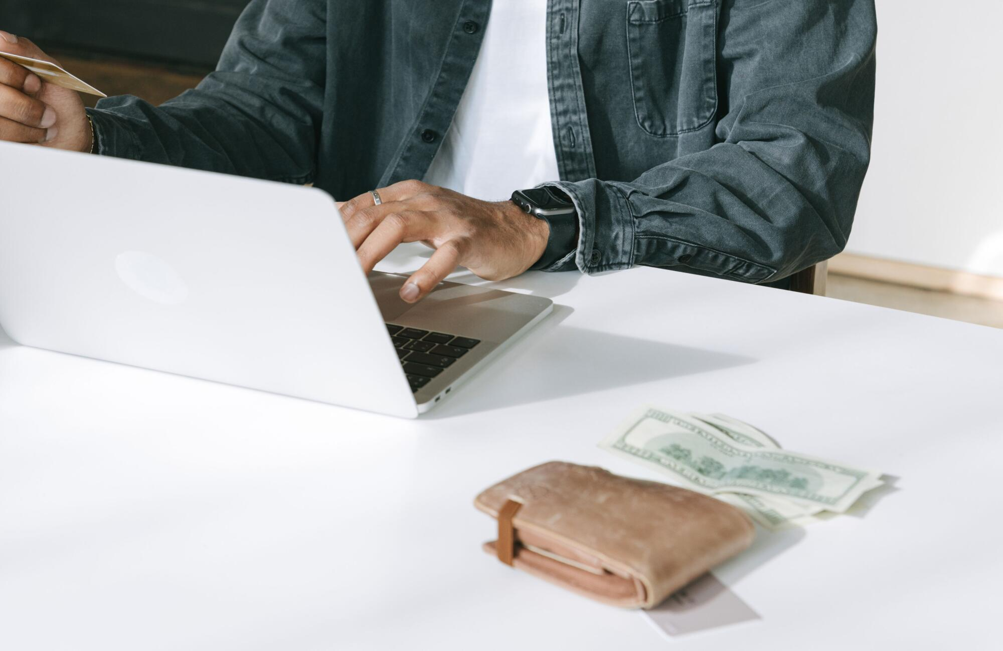 A man sits at a white table, working on a laptop and researching how to get an instant $100 cash advance online, with a wallet and cash out beside him.
