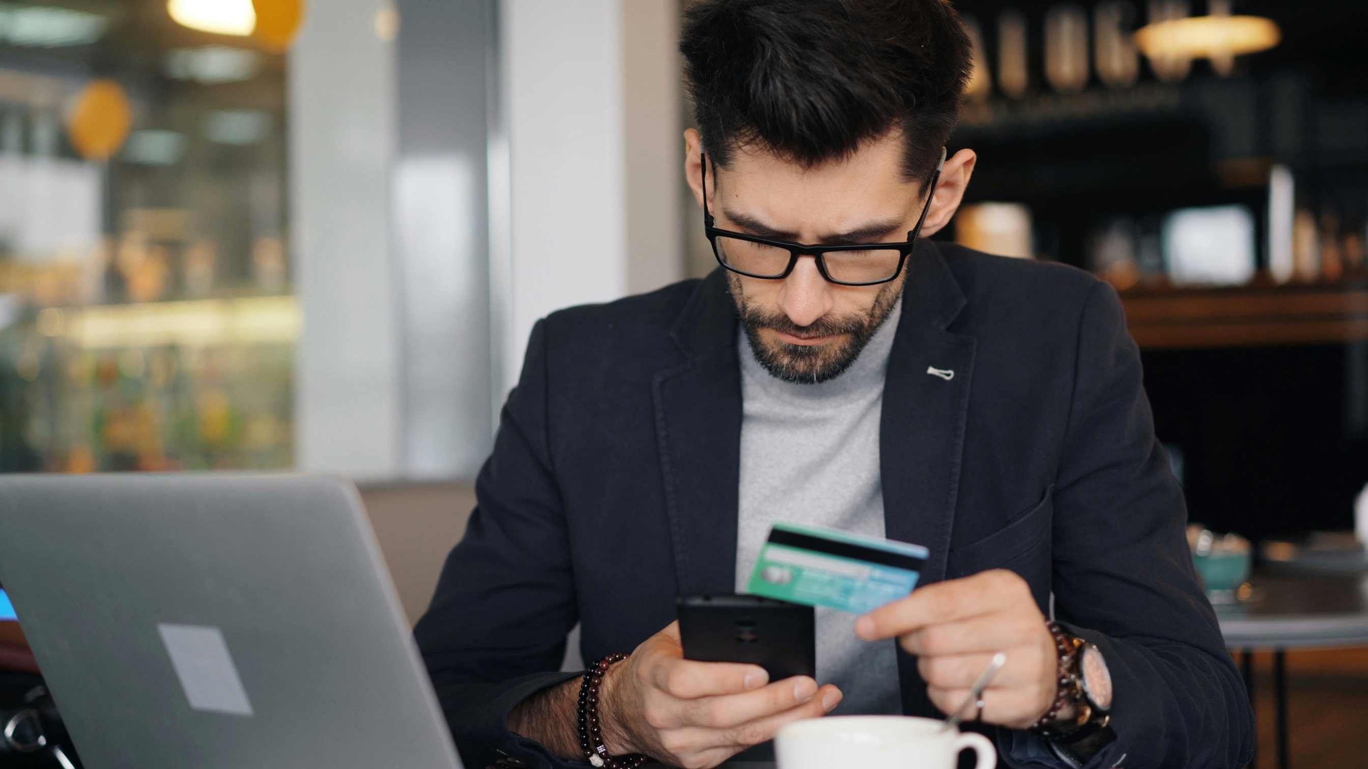 A person sitting at a table looking at his cell phone and holding a credit card