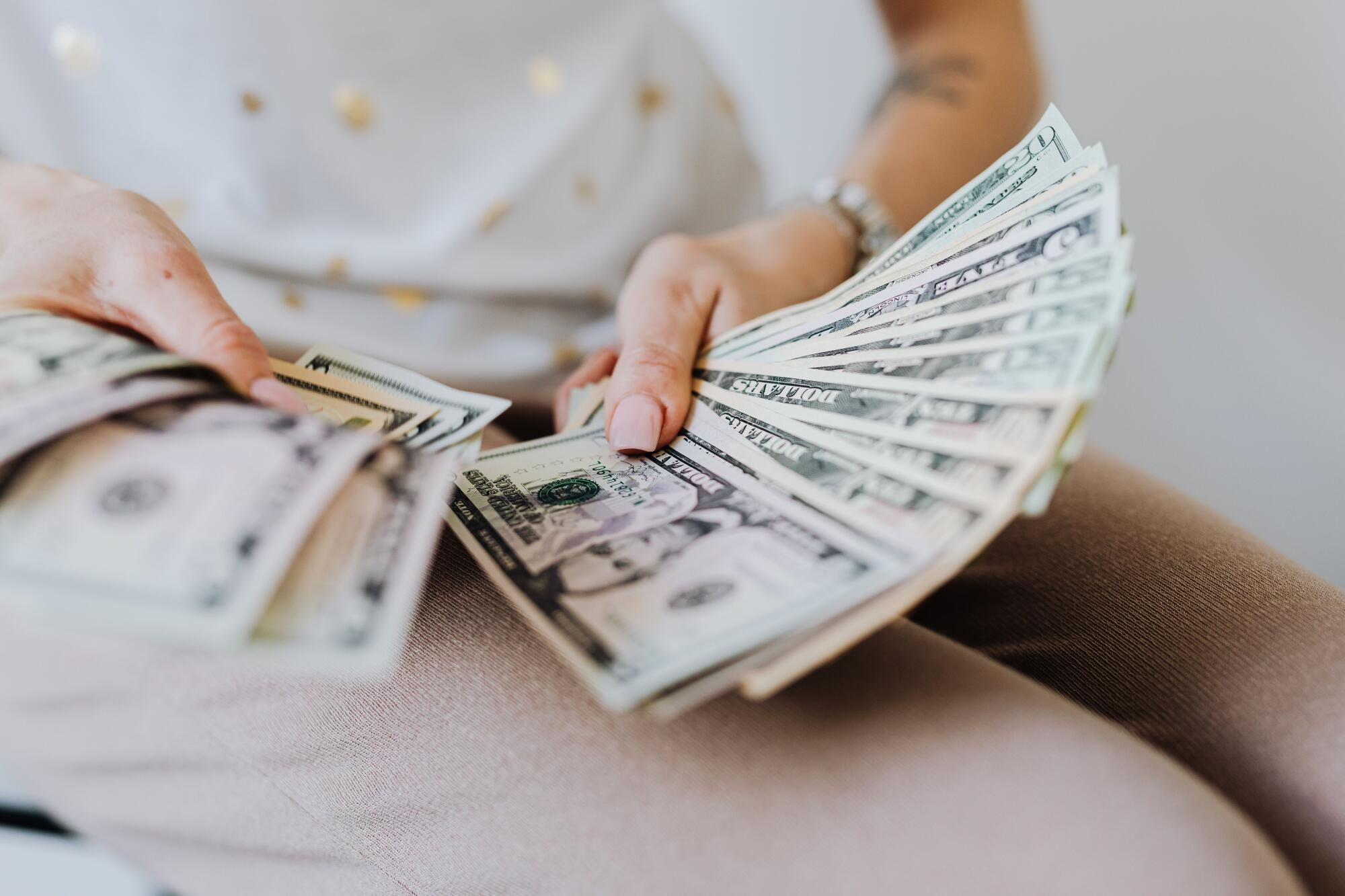 Image of a woman sitting down wearing light brown pants and a white shirt with gold polka dots who is holding a spread of American dollars representing a payout from payday loan lenders.